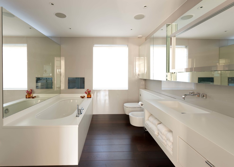 Inspiration pour une salle de bain minimaliste avec un placard à porte plane, des portes de placard blanches, WC suspendus, un mur blanc, parquet foncé, un lavabo intégré, un plan de toilette en surface solide, un sol marron et un plan de toilette blanc.