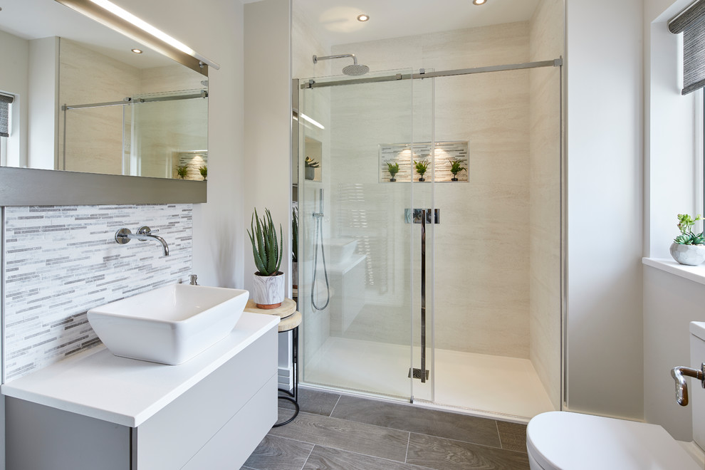 Exempel på ett mellanstort modernt vit vitt en-suite badrum, med en kantlös dusch, en toalettstol med hel cisternkåpa, grå kakel, porslinskakel, grå väggar, klinkergolv i porslin, ett väggmonterat handfat, bänkskiva i akrylsten, brunt golv och dusch med skjutdörr