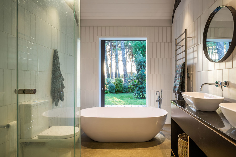 Inspiration for a contemporary ensuite bathroom in Auckland with flat-panel cabinets, black cabinets, a freestanding bath, white tiles, white walls, a vessel sink, beige floors, a hinged door and black worktops.