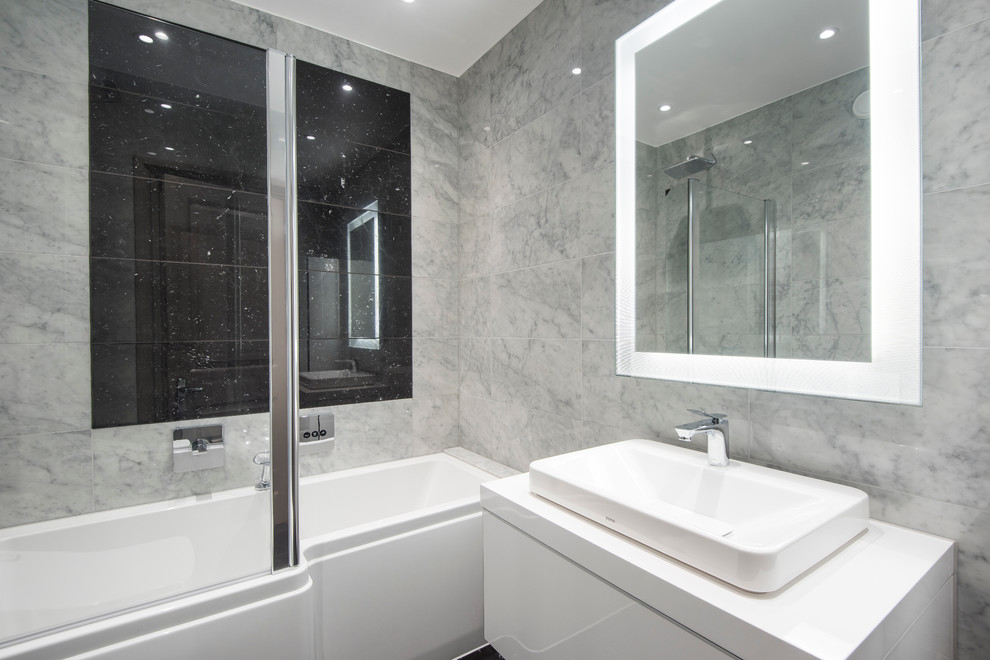 This is an example of a medium sized contemporary ensuite bathroom in London with flat-panel cabinets, white cabinets, an alcove bath, a shower/bath combination, a wall mounted toilet, grey tiles, stone tiles, grey walls, marble flooring and a vessel sink.