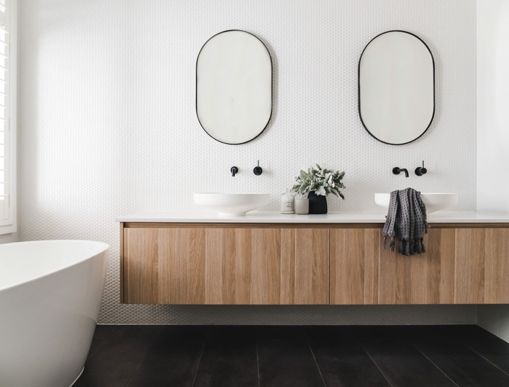 Inspiration for a contemporary ensuite bathroom in Melbourne with flat-panel cabinets, light wood cabinets, a freestanding bath, white tiles, mosaic tiles, white walls, a vessel sink, black floors, white worktops and double sinks.