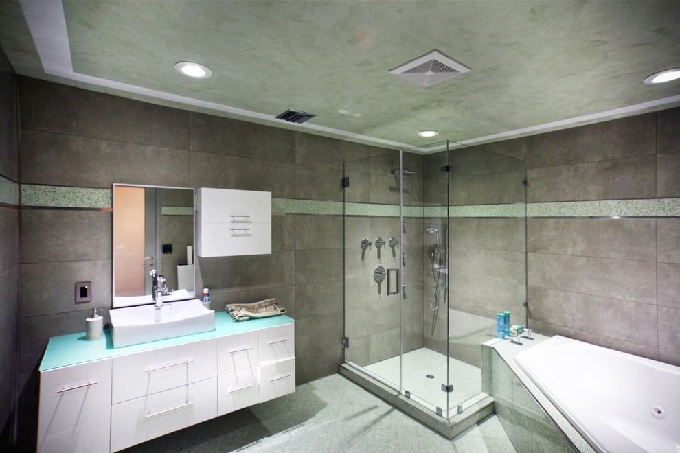Cette photo montre une grande salle de bain principale moderne avec un placard à porte plane, des portes de placard blanches, une baignoire d'angle, une douche d'angle, un carrelage gris, des carreaux de béton, un mur gris, un sol en carrelage de céramique, une vasque et un plan de toilette en verre.