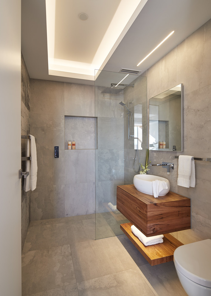 Foto på ett mellanstort funkis brun badrum med dusch, med släta luckor, skåp i mellenmörkt trä, en kantlös dusch, en vägghängd toalettstol, grå kakel, ett fristående handfat, träbänkskiva, grått golv och med dusch som är öppen