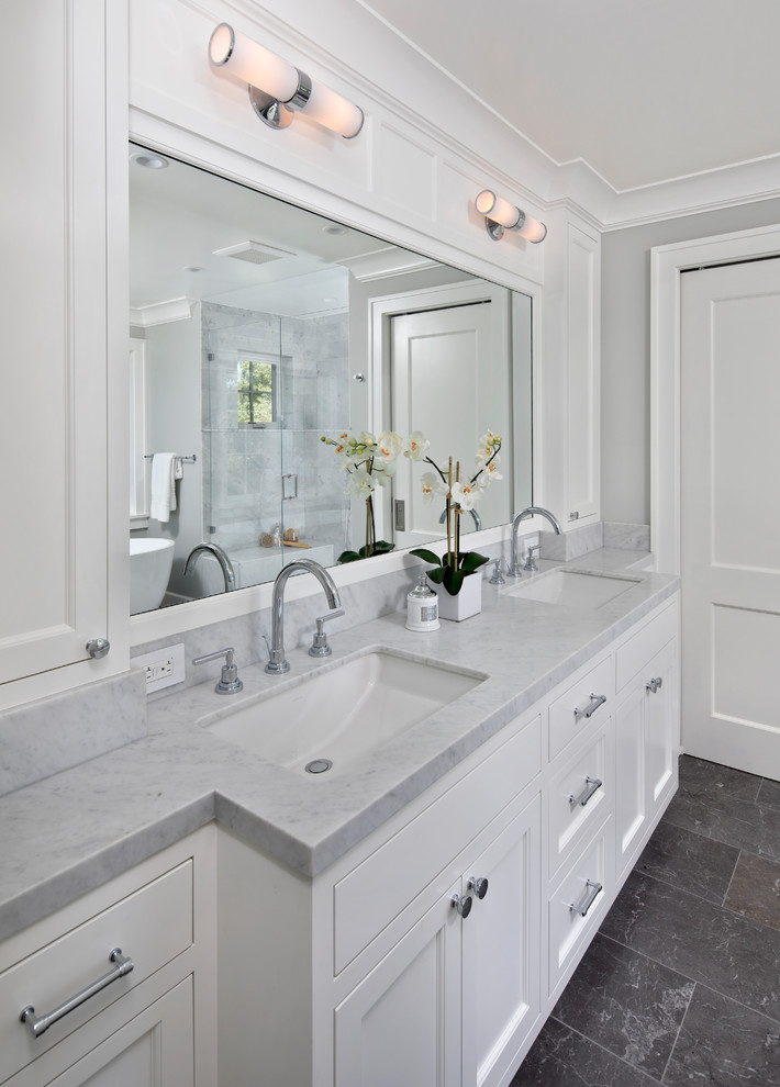 Пример оригинального дизайна: большая главная ванная комната в стиле неоклассика (современная классика) с фасадами с утопленной филенкой, белыми фасадами, унитазом-моноблоком, белой плиткой, мраморной плиткой, серыми стенами, полом из сланца, врезной раковиной, мраморной столешницей, черным полом, белой столешницей, отдельно стоящей ванной, угловым душем и душем с распашными дверями