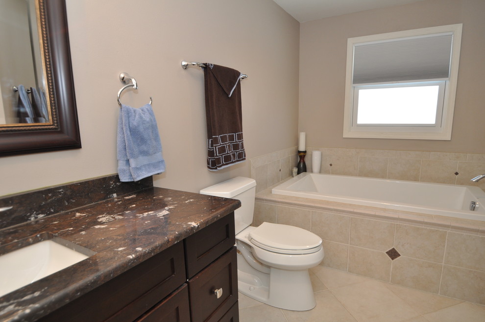 Example of a mid-sized master beige tile and ceramic tile ceramic tile drop-in bathtub design in Milwaukee with an undermount sink, raised-panel cabinets, dark wood cabinets, granite countertops, a two-piece toilet and gray walls
