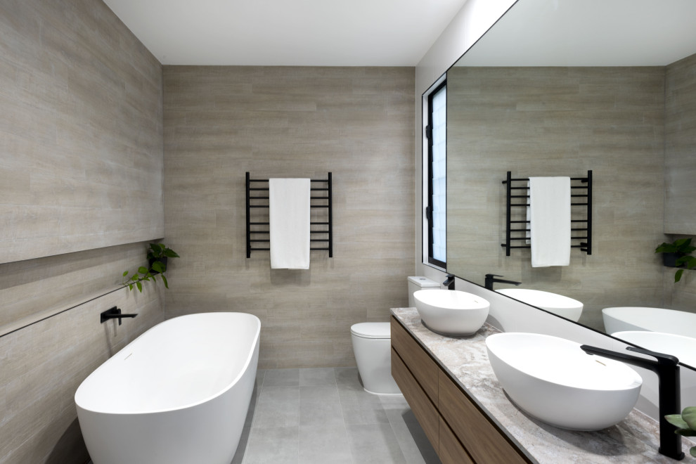 Inredning av ett modernt grå grått badrum, med släta luckor, skåp i mellenmörkt trä, ett fristående badkar, grå kakel, ett fristående handfat och grått golv