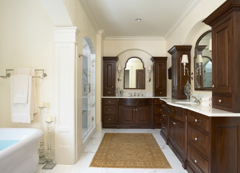 Klassisk inredning av ett vit vitt badrum, med ett undermonterad handfat, luckor med upphöjd panel, skåp i mörkt trä, marmorbänkskiva, en dusch i en alkov, en toalettstol med separat cisternkåpa, vit kakel och stenkakel