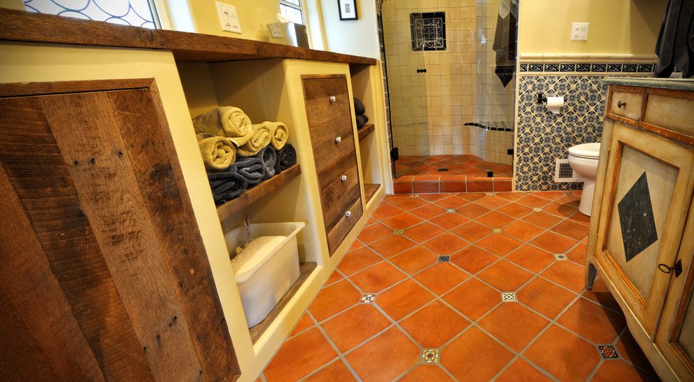 Réalisation d'une salle de bain sud-ouest américain en bois vieilli de taille moyenne avec un placard avec porte à panneau encastré, un mur jaune, tomettes au sol, une vasque, un plan de toilette en bois, un sol rouge et une cabine de douche à porte battante.