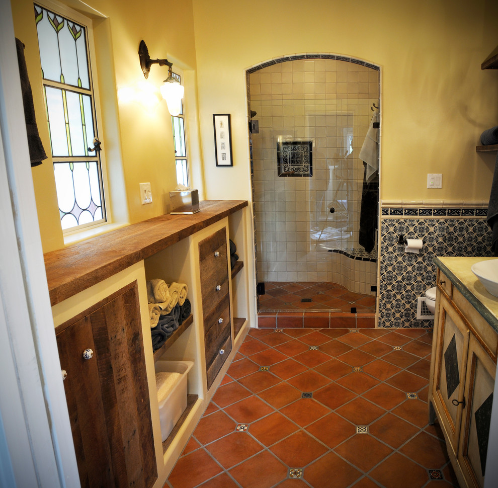 Inredning av ett amerikanskt mellanstort badrum med dusch, med klinkergolv i terrakotta, ett fristående handfat, rött golv, luckor med infälld panel, skåp i slitet trä, en dusch i en alkov, gula väggar, träbänkskiva och dusch med gångjärnsdörr