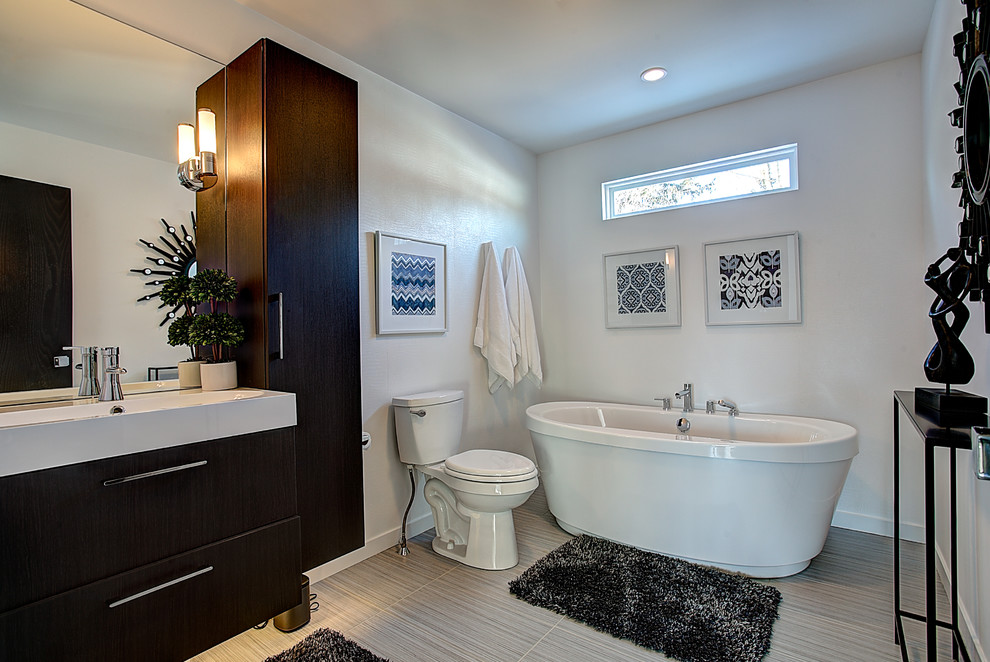 Modernes Badezimmer mit flächenbündigen Schrankfronten, dunklen Holzschränken, Mineralwerkstoff-Waschtisch, freistehender Badewanne, Keramikfliesen, weißer Wandfarbe, Keramikboden und Trogwaschbecken in Grand Rapids