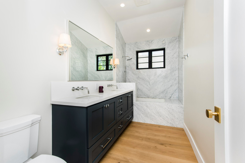 Design ideas for a medium sized traditional shower room bathroom in Los Angeles with shaker cabinets, blue cabinets, a submerged bath, a shower/bath combination, a one-piece toilet, black and white tiles, marble tiles, white walls, light hardwood flooring, a submerged sink, engineered stone worktops, brown floors, an open shower, white worktops, double sinks and a built in vanity unit.