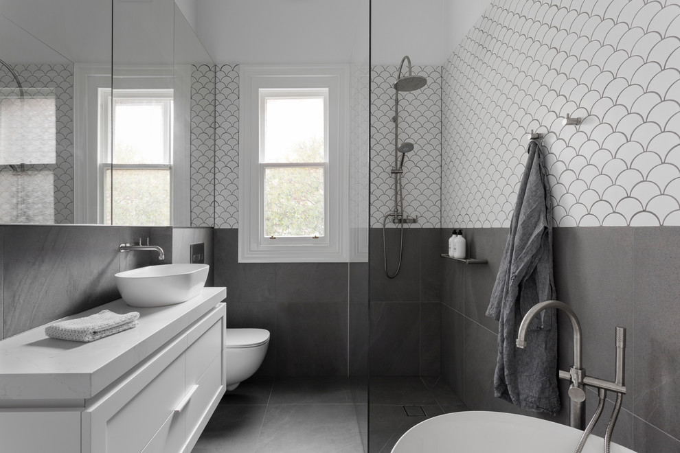 Modern inredning av ett vit vitt badrum med dusch, med vita skåp, ett fristående badkar, våtrum, en vägghängd toalettstol, grå kakel, vit kakel, vita väggar, ett fristående handfat, grått golv och med dusch som är öppen