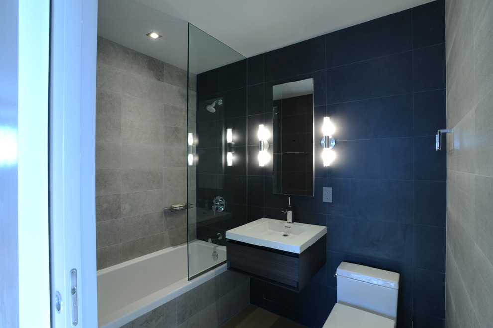 Exempel på ett mellanstort modernt badrum, med ett väggmonterat handfat, skåp i mörkt trä, bänkskiva i akrylsten, ett badkar i en alkov, en dusch/badkar-kombination, en toalettstol med hel cisternkåpa, svart kakel, stenkakel, grå väggar och ljust trägolv