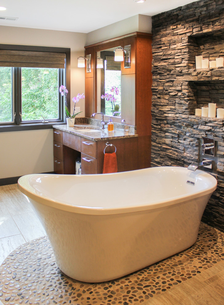 Cette image montre une salle de bain principale design en bois brun de taille moyenne avec un placard à porte plane, un plan de toilette en granite, une baignoire indépendante, un carrelage marron, un carrelage de pierre, un mur beige et un lavabo encastré.