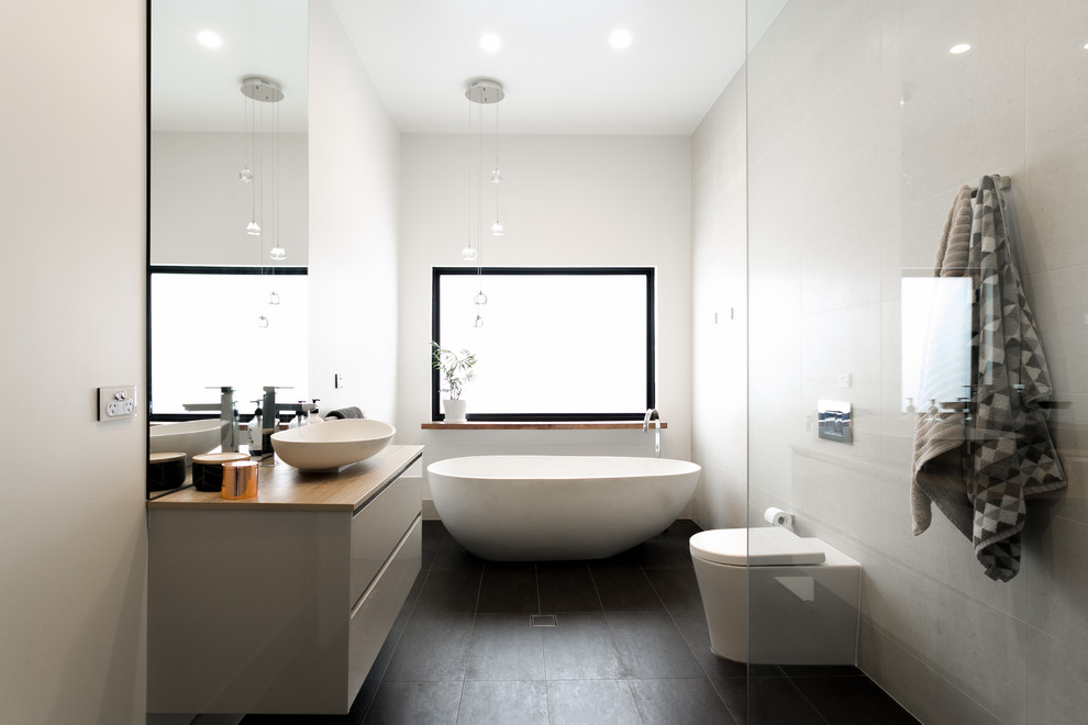 Bild på ett funkis beige beige en-suite badrum, med släta luckor, beige skåp, ett fristående badkar, en öppen dusch, en toalettstol med hel cisternkåpa, vit kakel, ett fristående handfat, träbänkskiva, svart golv och med dusch som är öppen