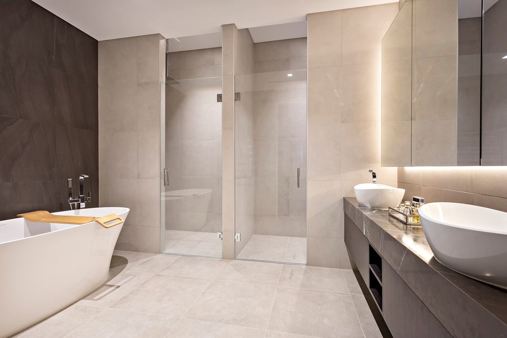 Cette image montre une salle de bain design avec un placard à porte plane, des portes de placard grises, une baignoire indépendante, une douche à l'italienne, un carrelage beige, une vasque, un sol beige et un plan de toilette gris.