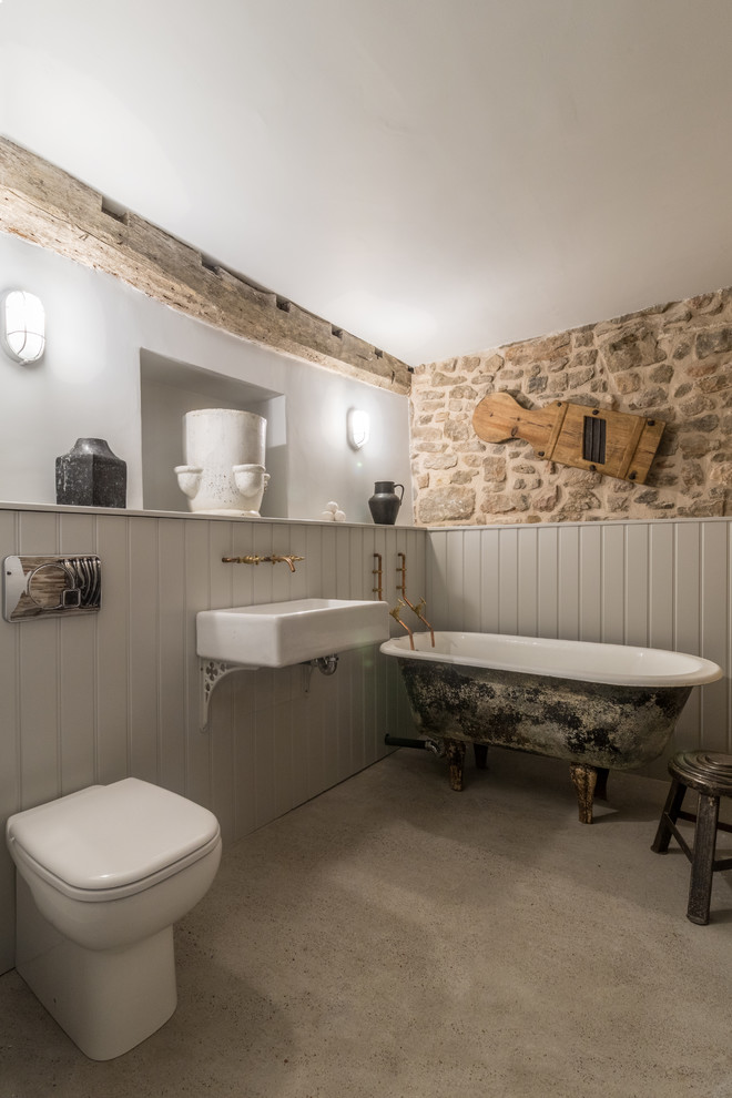 Modelo de cuarto de baño campestre de tamaño medio con bañera exenta, sanitario de una pieza, paredes grises, suelo de cemento, lavabo suspendido y piedra
