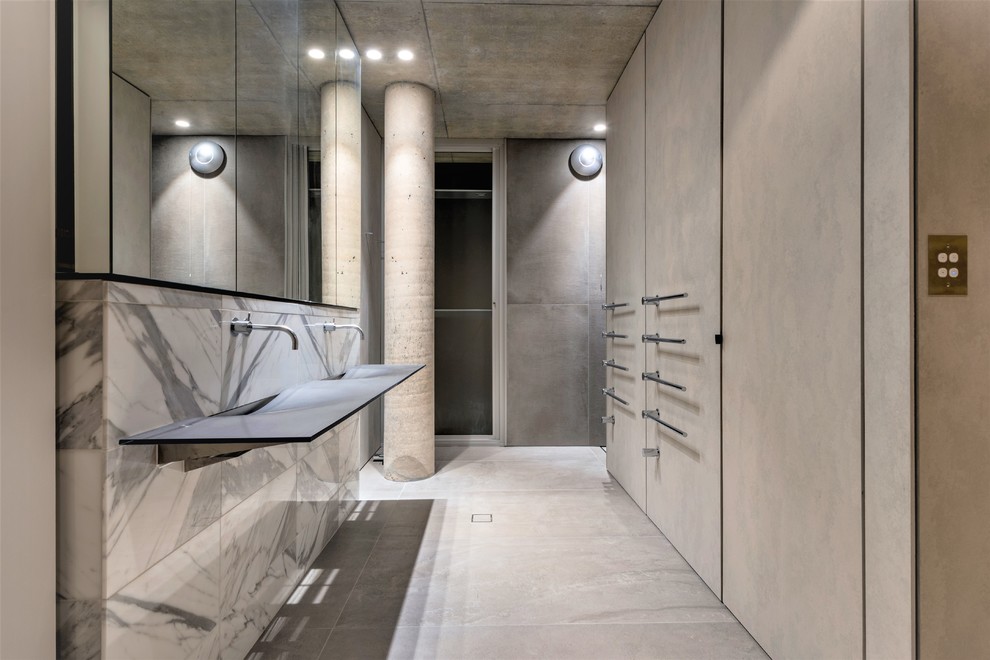 Industriell inredning av ett en-suite badrum, med grå kakel, vit kakel, grå väggar, betonggolv, ett väggmonterat handfat, grått golv, luckor med glaspanel, travertinkakel och bänkskiva i glas