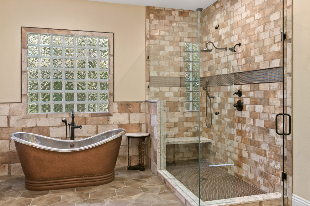 На фото: большая главная ванная комната в средиземноморском стиле с отдельно стоящей ванной, угловым душем, бежевыми стенами, серым полом, душем с распашными дверями, сиденьем для душа и кирпичными стенами с