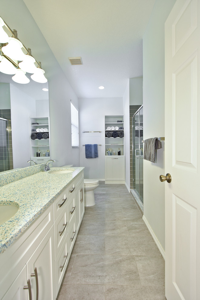 タンパにあるお手頃価格の中くらいなトランジショナルスタイルのおしゃれなマスターバスルーム (シェーカースタイル扉のキャビネット、白いキャビネット、グレーのタイル、サブウェイタイル、磁器タイルの床、アンダーカウンター洗面器、再生グラスカウンター、アルコーブ型シャワー、分離型トイレ、白い壁、グレーの床、引戸のシャワー) の写真