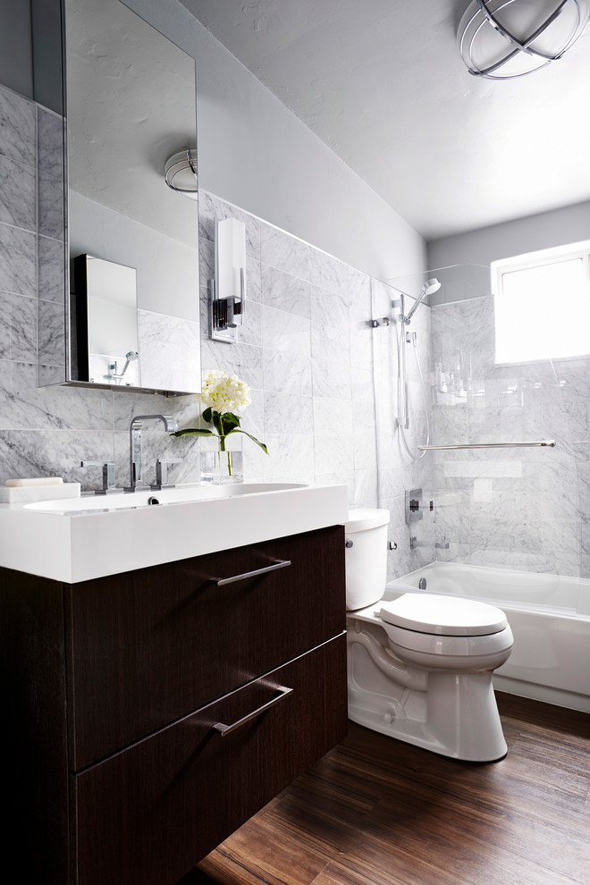 Bild på ett litet funkis badrum, med ett integrerad handfat, släta luckor, skåp i mörkt trä, ett badkar i en alkov, en dusch/badkar-kombination, en toalettstol med separat cisternkåpa, grå kakel, stenkakel och grå väggar