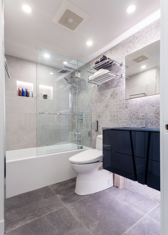 Идея дизайна: ванная комната среднего размера в современном стиле с плоскими фасадами, ванной в нише, душем над ванной, унитазом-моноблоком, серой плиткой, керамогранитной плиткой, серыми стенами, монолитной раковиной, стеклянной столешницей, серым полом, душем с распашными дверями, синей столешницей, черными фасадами, полом из цементной плитки и душевой кабиной