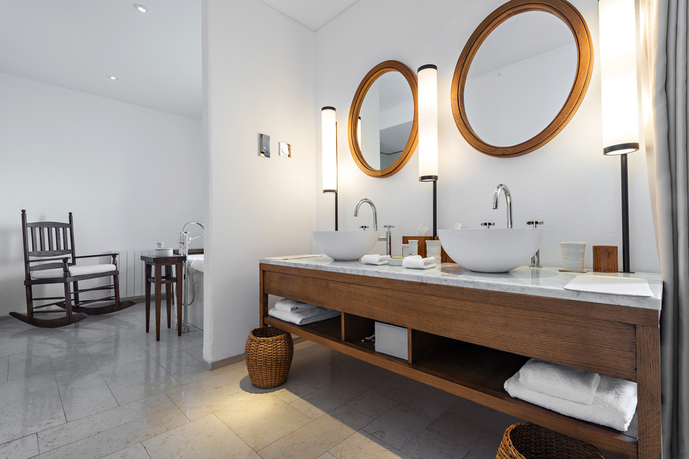 На фото: большая главная ванная комната в современном стиле с темными деревянными фасадами, бежевым полом, открытыми фасадами, отдельно стоящей ванной, белыми стенами, полом из известняка, настольной раковиной и мраморной столешницей с