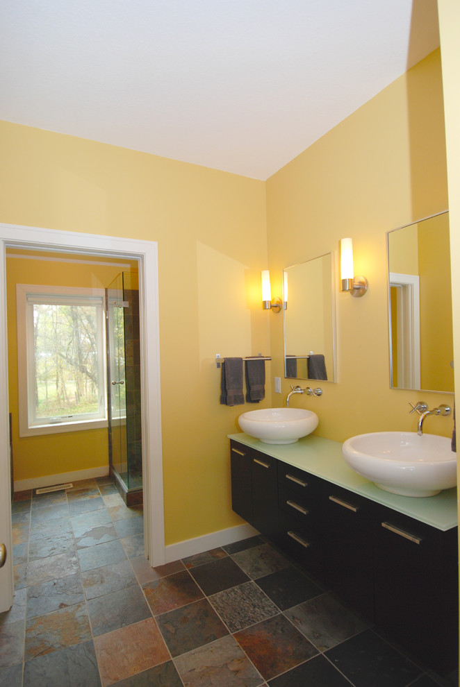 Bild på ett mellanstort funkis badrum, med ett väggmonterat handfat, släta luckor, skåp i mörkt trä, bänkskiva i glas, en hörndusch, en toalettstol med separat cisternkåpa, brun kakel, stenkakel, gula väggar och skiffergolv