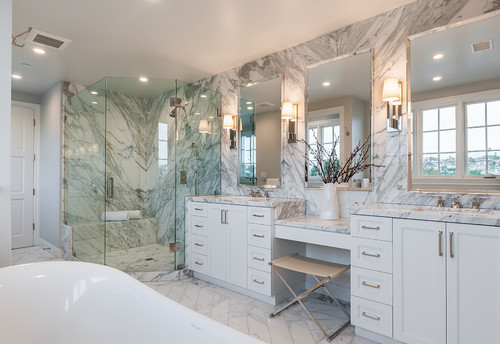 Elegant bathroom with white double vanities, marble walls, glass shower enclosure, and built-in makeup station