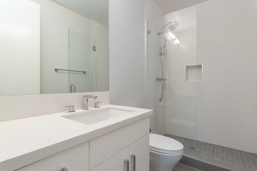 Пример оригинального дизайна: большая ванная комната в стиле модернизм с плоскими фасадами, белыми фасадами, душем в нише, унитазом-моноблоком, белой плиткой, керамогранитной плиткой, серыми стенами, полом из керамогранита, душевой кабиной, врезной раковиной, столешницей из искусственного кварца, серым полом, душем с распашными дверями и белой столешницей