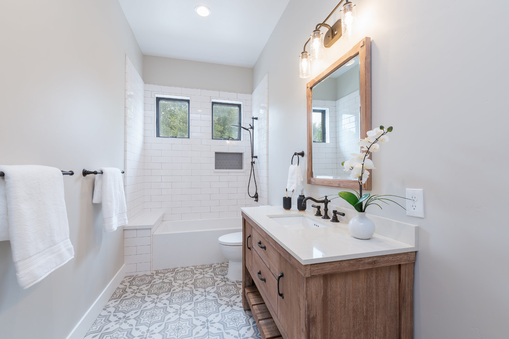 Bild på ett mellanstort lantligt vit vitt badrum med dusch, med bruna skåp, ett badkar i en alkov, en dusch/badkar-kombination, en toalettstol med hel cisternkåpa, grå kakel, keramikplattor, grå väggar, klinkergolv i keramik, ett undermonterad handfat, bänkskiva i kvartsit, grått golv, dusch med duschdraperi och släta luckor