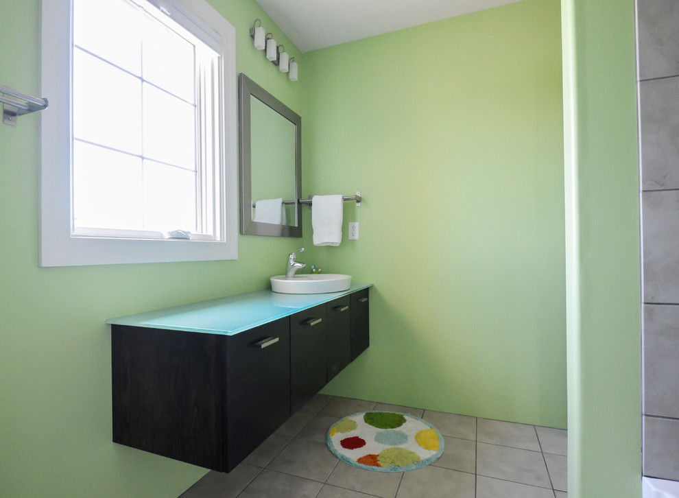 Immagine di una grande stanza da bagno per bambini minimalista con lavabo a bacinella, ante lisce, ante in legno bruno, top in vetro, vasca/doccia, piastrelle grigie, piastrelle di cemento, pareti verdi e pavimento con piastrelle in ceramica
