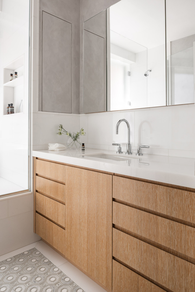 Modernes Badezimmer mit flächenbündigen Schrankfronten, hellbraunen Holzschränken, weißen Fliesen, Unterbauwaschbecken, grauem Boden und weißer Waschtischplatte in New York