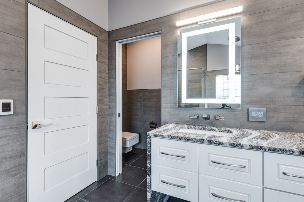 This is an example of a large modern ensuite bathroom in Chicago with raised-panel cabinets, white cabinets, a freestanding bath, a walk-in shower, a wall mounted toilet, grey tiles, ceramic tiles, grey walls, ceramic flooring, a submerged sink, quartz worktops, grey floors and a hinged door.