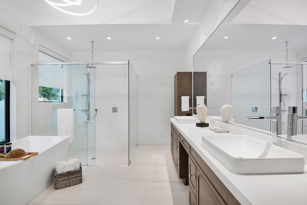 Photo of a medium sized contemporary ensuite bathroom in Miami with flat-panel cabinets, light wood cabinets, a freestanding bath, a walk-in shower, a one-piece toilet, grey tiles, porcelain tiles, white walls, porcelain flooring, a vessel sink, engineered stone worktops, white floors, a sliding door, white worktops, an enclosed toilet, double sinks, a freestanding vanity unit and a drop ceiling.