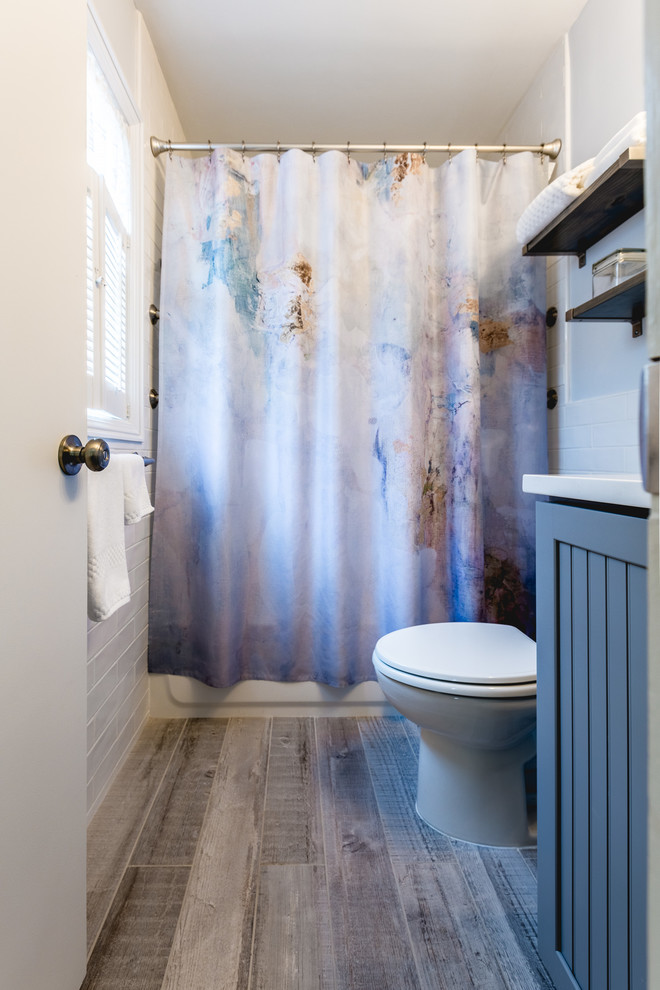 Idéer för ett mellanstort lantligt vit badrum, med luckor med lamellpanel, en toalettstol med hel cisternkåpa, grå skåp, ett badkar i en alkov, en dusch/badkar-kombination, vit kakel, blå väggar, mörkt trägolv, ett undermonterad handfat, brunt golv och dusch med duschdraperi