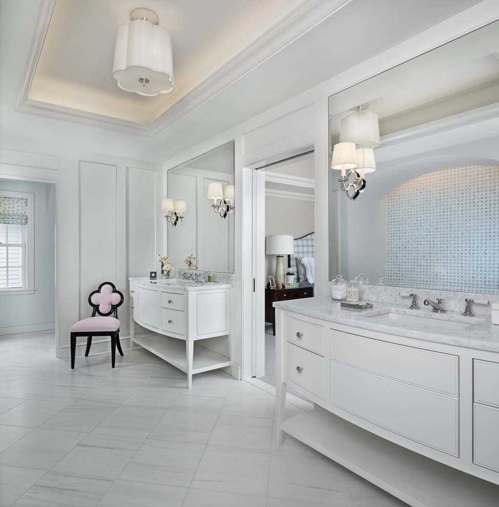 На фото: огромная главная ванная комната в классическом стиле с белыми фасадами, белыми стенами, полом из травертина, врезной раковиной, мраморной столешницей, белым полом, белой столешницей, зеркалом с подсветкой и плоскими фасадами