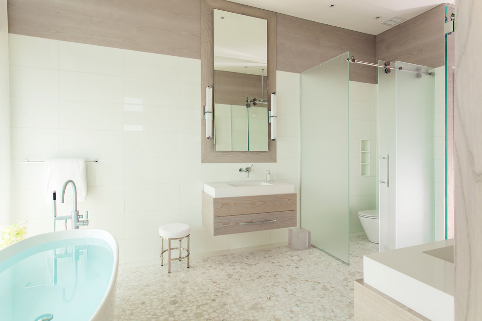 Пример оригинального дизайна: большая главная ванная комната в современном стиле с отдельно стоящей ванной, бежевой плиткой, подвесной раковиной, раздельным унитазом, плиткой мозаикой, бежевыми стенами, плоскими фасадами, светлыми деревянными фасадами, душем в нише, полом из мозаичной плитки, столешницей из искусственного камня, бежевым полом, душем с раздвижными дверями и белой столешницей