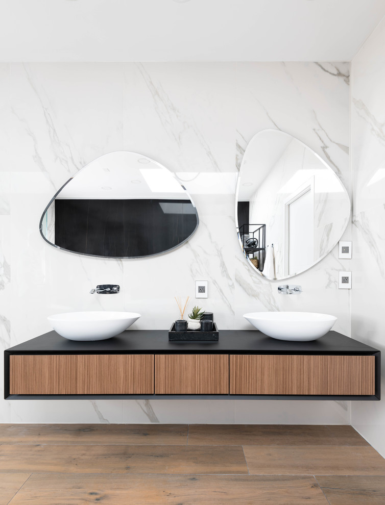 This is an example of a large contemporary ensuite bathroom in Vancouver with flat-panel cabinets, medium wood cabinets, a freestanding bath, a one-piece toilet, white tiles, white walls, a vessel sink, brown floors, a hinged door, black worktops, a corner shower, stone slabs, medium hardwood flooring and solid surface worktops.