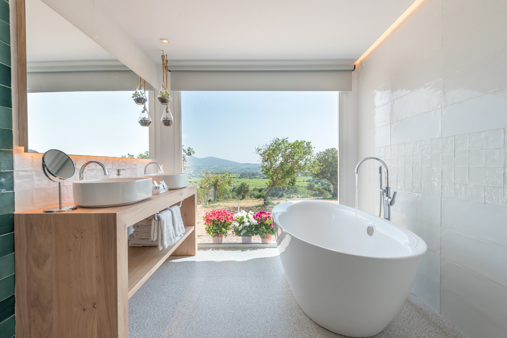 На фото: главная ванная комната в современном стиле с открытыми фасадами, светлыми деревянными фасадами, отдельно стоящей ванной, белой плиткой, настольной раковиной, столешницей из дерева, серым полом и коричневой столешницей
