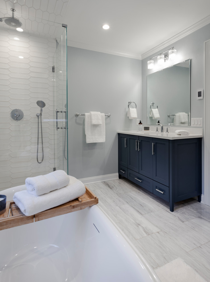 Photo of a large modern ensuite bathroom in New York with beaded cabinets, blue cabinets, a freestanding bath, a corner shower, a one-piece toilet, white tiles, ceramic tiles, grey walls, marble flooring, a submerged sink, solid surface worktops, grey floors, a hinged door, white worktops, a wall niche, double sinks and a built in vanity unit.