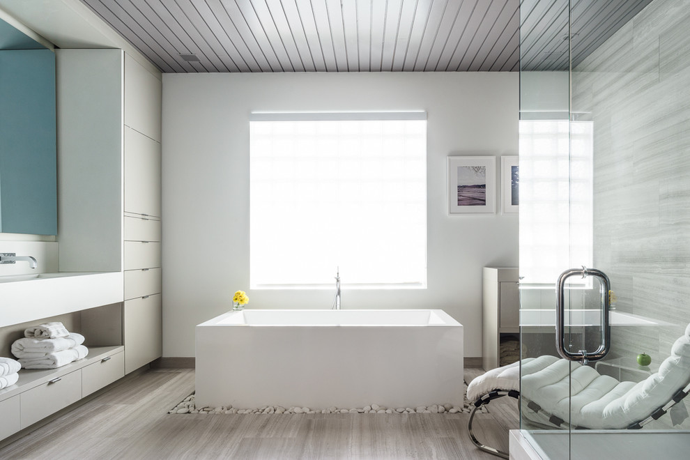 На фото: большая главная ванная комната в современном стиле с монолитной раковиной, плоскими фасадами, серыми фасадами, отдельно стоящей ванной, белыми стенами и каменной плиткой