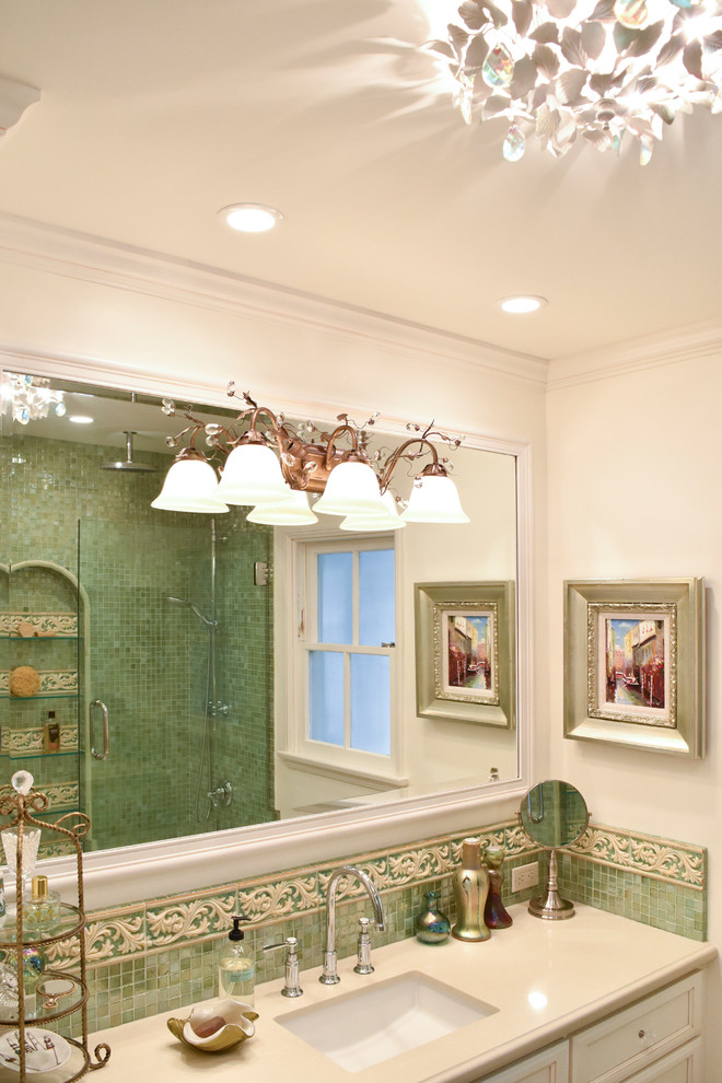 На фото: большая главная ванная комната в средиземноморском стиле с врезной раковиной, фасадами с утопленной филенкой, искусственно-состаренными фасадами, столешницей из искусственного камня, угловой ванной, душем над ванной, унитазом-моноблоком, зеленой плиткой, стеклянной плиткой, бежевыми стенами и полом из керамогранита с