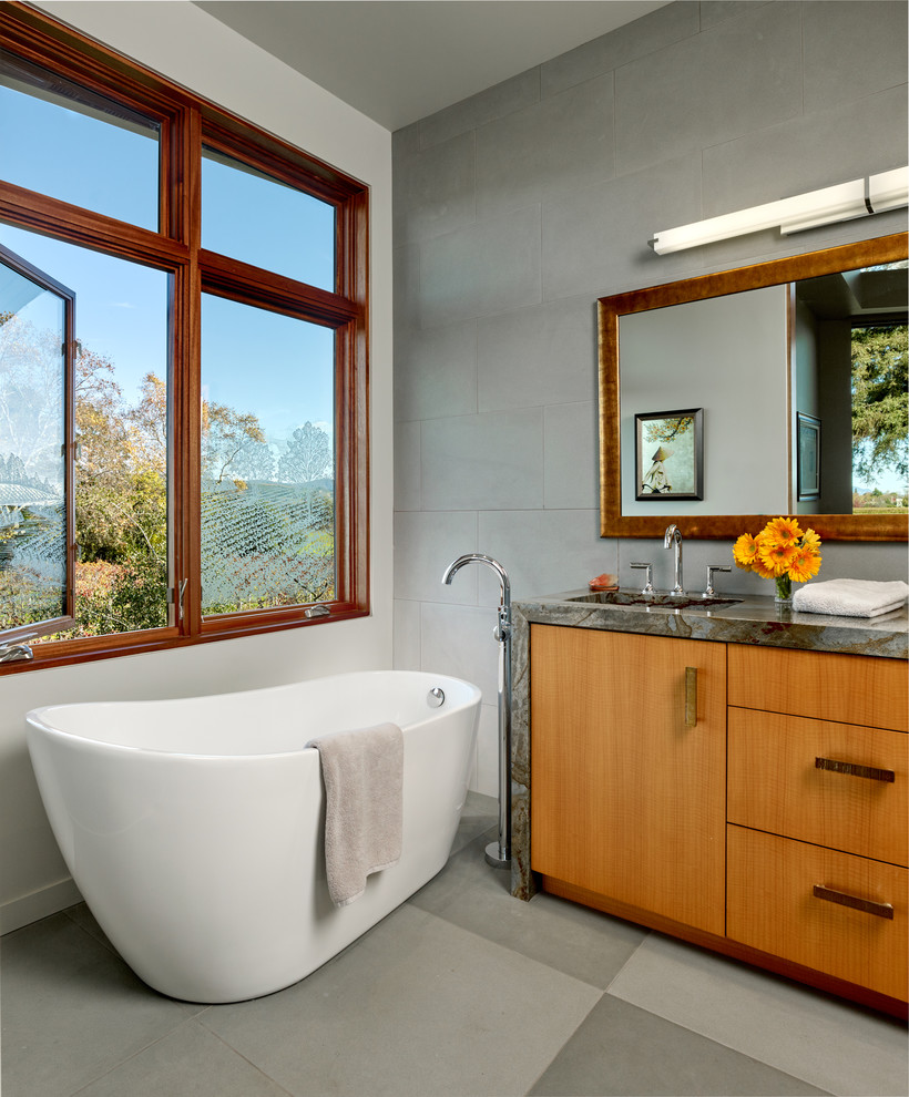 Inredning av ett modernt mellanstort en-suite badrum, med släta luckor, skåp i ljust trä, ett fristående badkar, granitbänkskiva, grå väggar, ett undermonterad handfat, grå kakel och keramikplattor