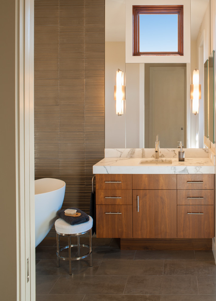 Ejemplo de cuarto de baño minimalista con lavabo bajoencimera, armarios con paneles lisos, puertas de armario de madera oscura, encimera de mármol, bañera exenta y baldosas y/o azulejos grises