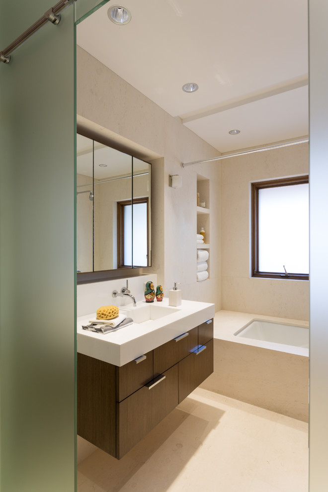Cette photo montre une salle de bain moderne en bois foncé avec un lavabo intégré, un placard à porte plane, une baignoire encastrée, un combiné douche/baignoire et un carrelage beige.