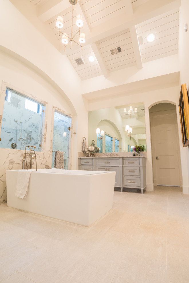 Свежая идея для дизайна: большая главная ванная комната в классическом стиле с фасадами с утопленной филенкой, серыми фасадами, отдельно стоящей ванной, двойным душем, бежевой плиткой, мраморной плиткой, белыми стенами, полом из керамической плитки, врезной раковиной, мраморной столешницей, бежевым полом, душем с распашными дверями и бежевой столешницей - отличное фото интерьера