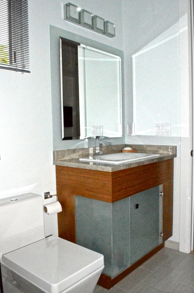 Inredning av ett modernt litet badrum, med ett nedsänkt handfat, luckor med glaspanel, skåp i mellenmörkt trä, granitbänkskiva, ett badkar i en alkov, en dusch/badkar-kombination, en toalettstol med hel cisternkåpa, grå kakel, porslinskakel, grå väggar och klinkergolv i porslin
