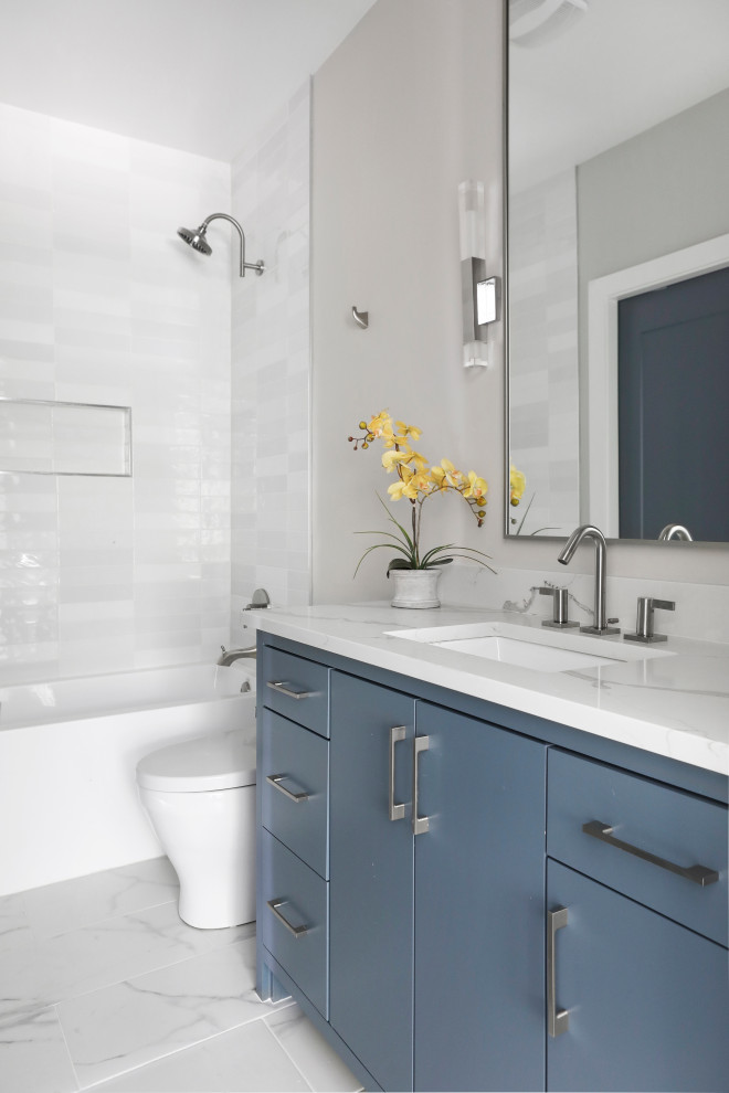 Cette image montre une salle de bain design avec un placard à porte plane, des portes de placard bleues, une baignoire en alcôve, un combiné douche/baignoire, WC à poser, un carrelage blanc, un carrelage métro, un mur gris, un sol en carrelage de porcelaine, un lavabo encastré, un plan de toilette en quartz modifié, un sol blanc, un plan de toilette blanc, une niche, meuble simple vasque et meuble-lavabo encastré.