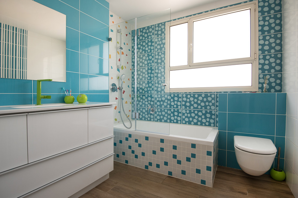 Idée de décoration pour une salle d'eau design avec un placard à porte plane, des portes de placard blanches, une baignoire posée, un combiné douche/baignoire, WC suspendus, un carrelage bleu, un mur bleu, un sol en bois brun, un lavabo encastré, un sol marron et aucune cabine.